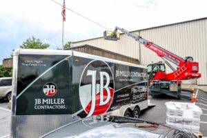 JB Miller Contractors trailer with logo, commercial roofing equipment in use, and American flag in background, emphasizing roofing expertise and services.